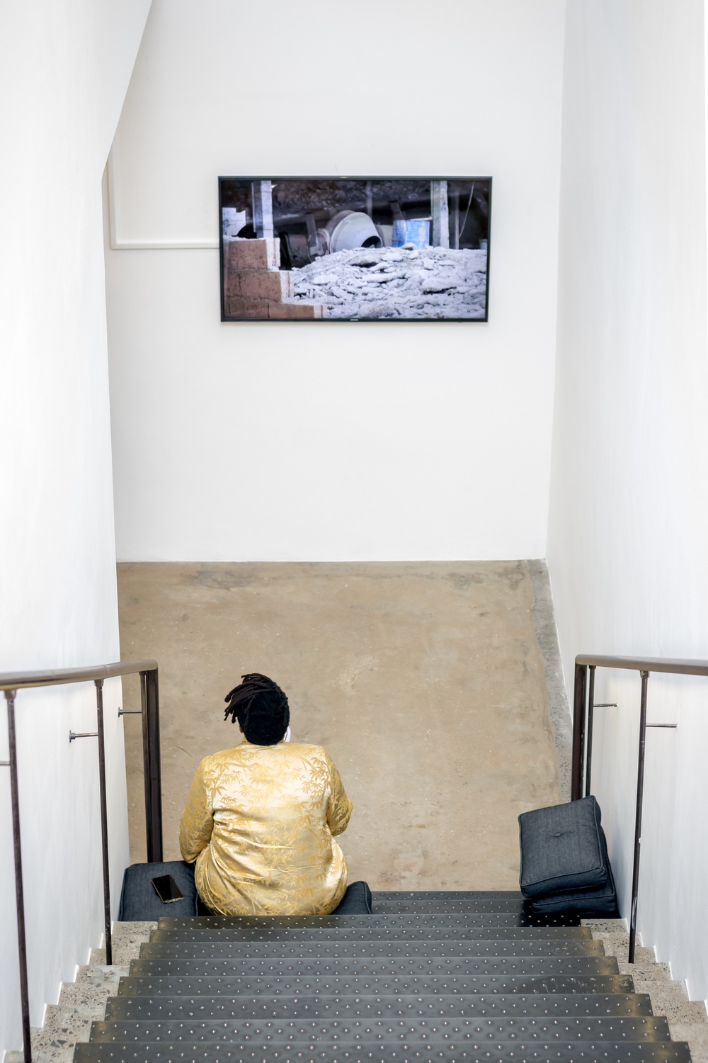 Event photograph from the opening of the You & I exhibition in A4’s Gallery. At the back, Adrian Melis’ video work is playing on a wall-mounted screen. At the front, an attendee sits on A4’s staircase.
