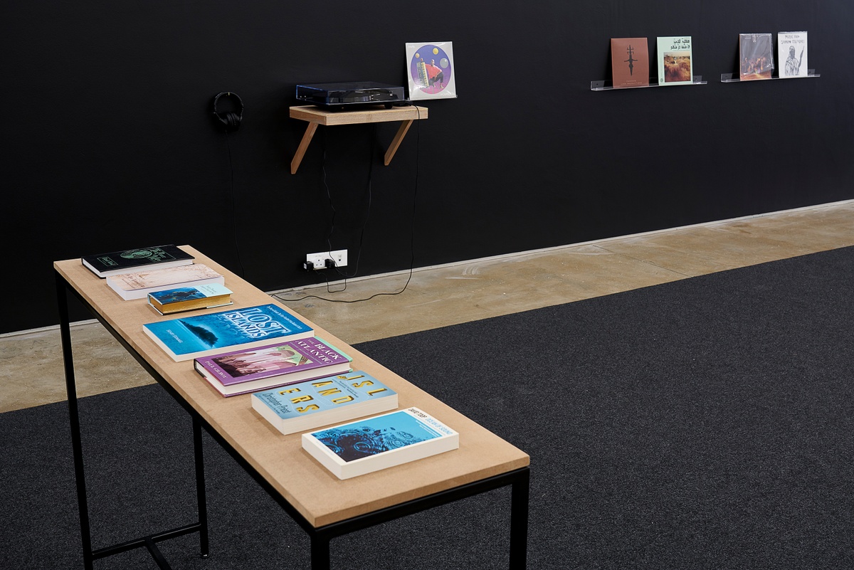 Installation photograph from the ‘Sounding the Void, Imaging the Orchestra V.1’ exhibition in A4’s Gallery. On the left, a table is laden with reading materials from Andrew Pekler’s installation ‘Phantom Islands - A Sonic Atlas’. In the middle, a listening station features a record player sitting on a wall-mounted shelf. On the right, a selection of vinyl records from the Sahel Sounds record label sits on wall-mounted shelves.
