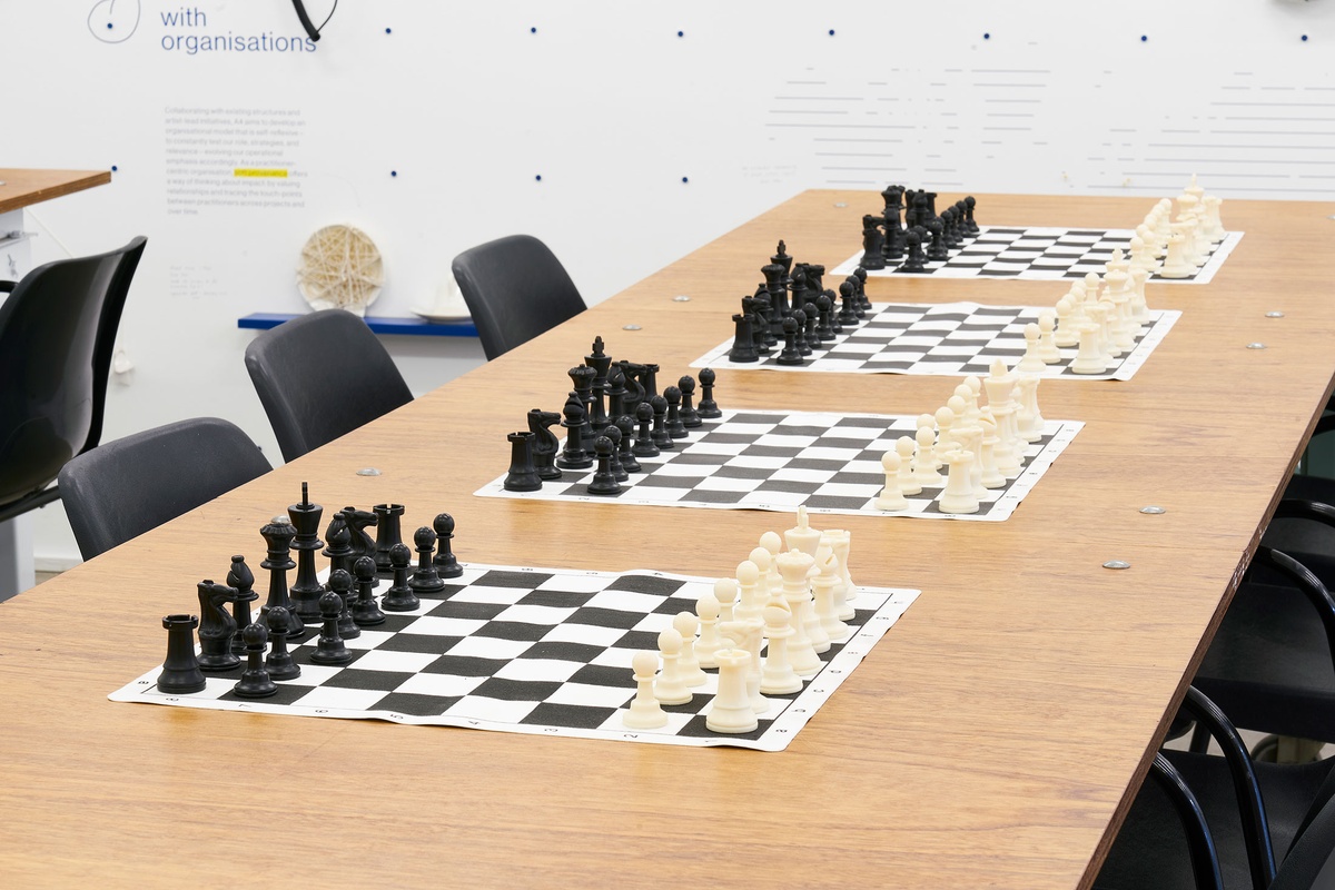 Installation photograph from the launch of Brett Seiler’s chess set in A4’s Proto museum shop that shows the chess tournament setup. A long wooden table with chairs host a series of travelling chess sets.
