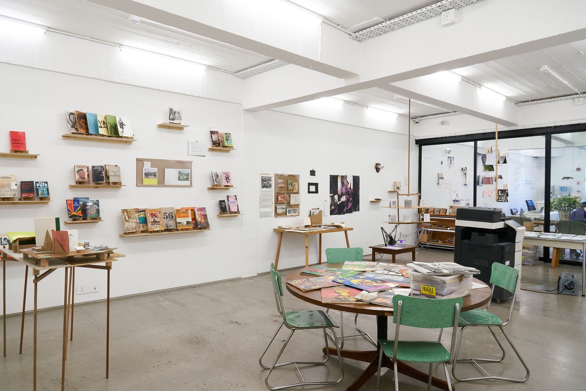 Installation photograph from the Papertrails exhibition in A4’s Reading Room. On the right, a round table strewn with printed matter. On the left, various wall-mounted shelves with printed matter.
