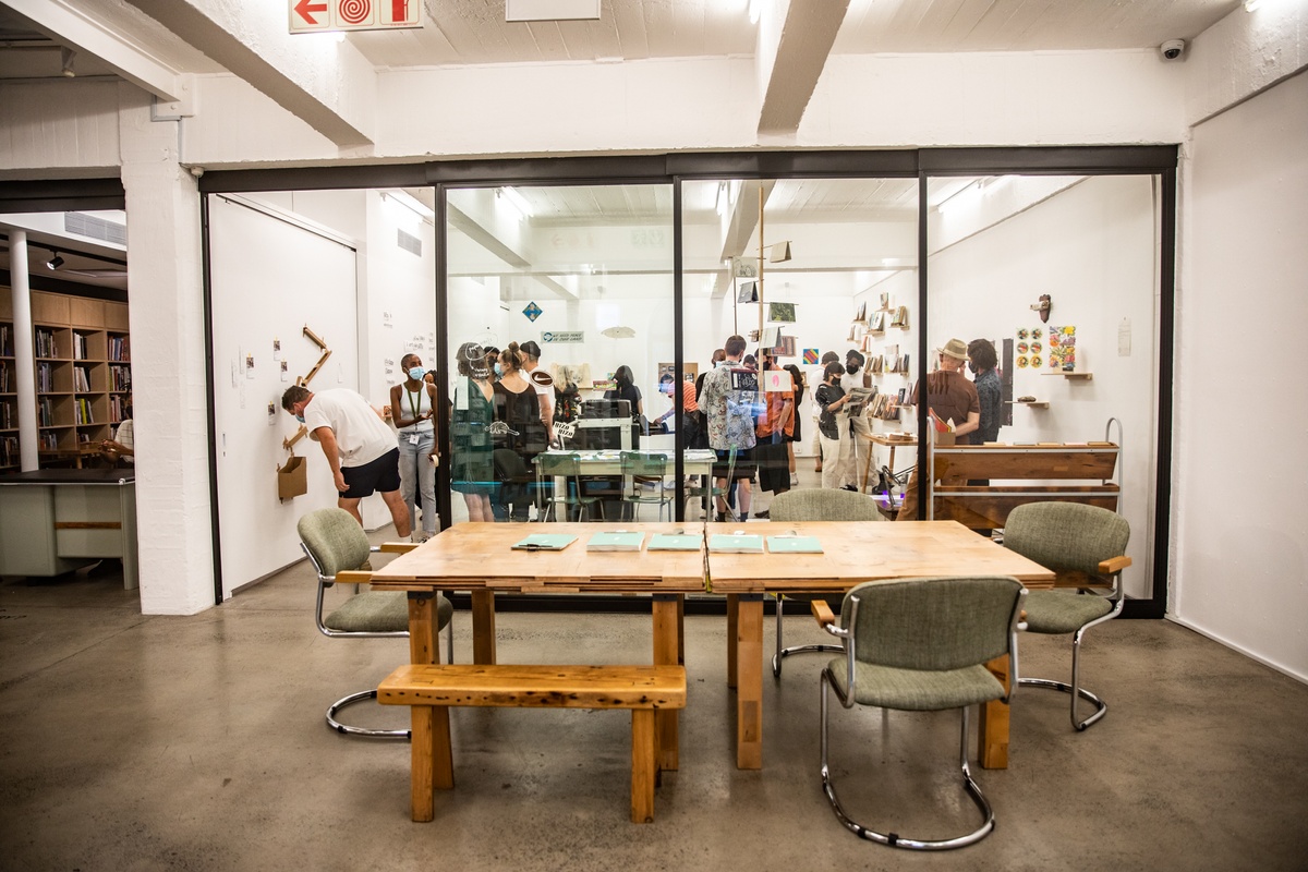 Event photograph from the opening of the Papertrails exhibition in A4’s Reading Room. At the front, two square wooden tables with seating. At the back, the Papertrails working studio filled with participants.
