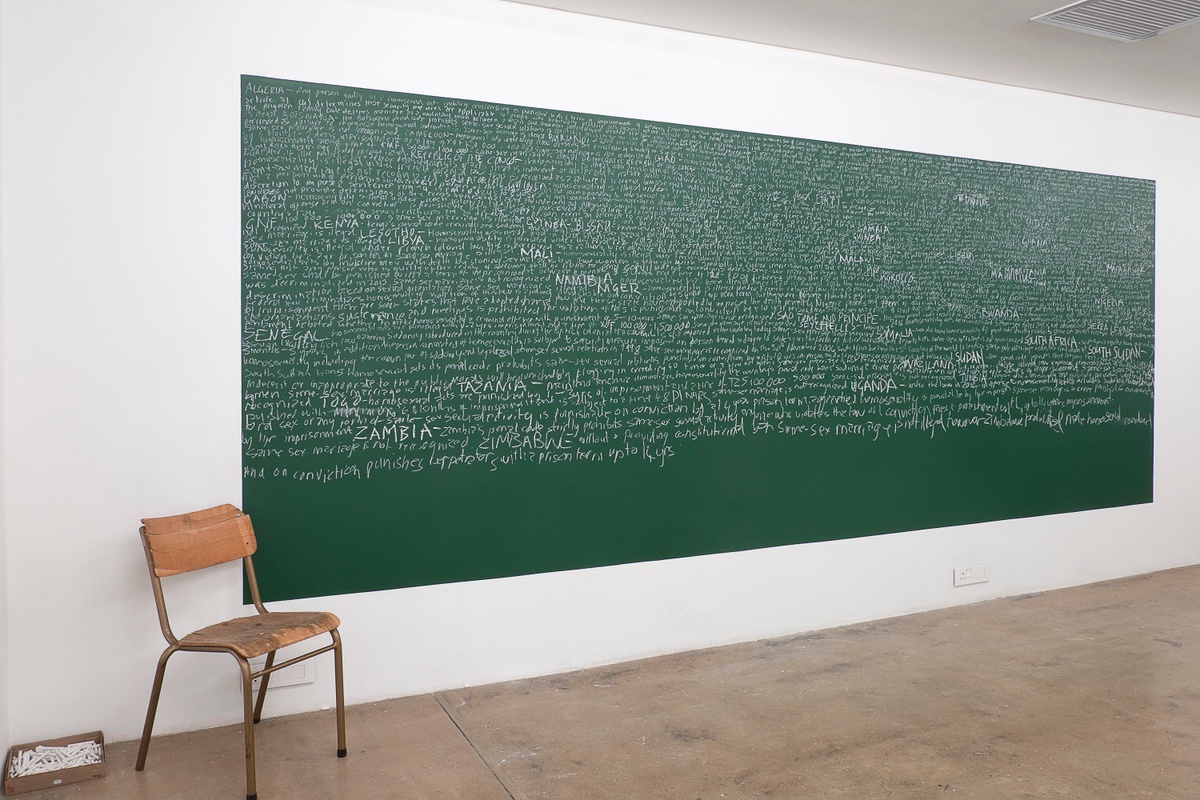 Installation photograph from the You & I exhibition in A4’s Gallery. At the back, a chalkboard with chalk writing from Brett Seiler and Luvuyo Nyawose’s performance installation ‘Reading Homophobia’ is mounted on the wall, with a single chair on the left.
