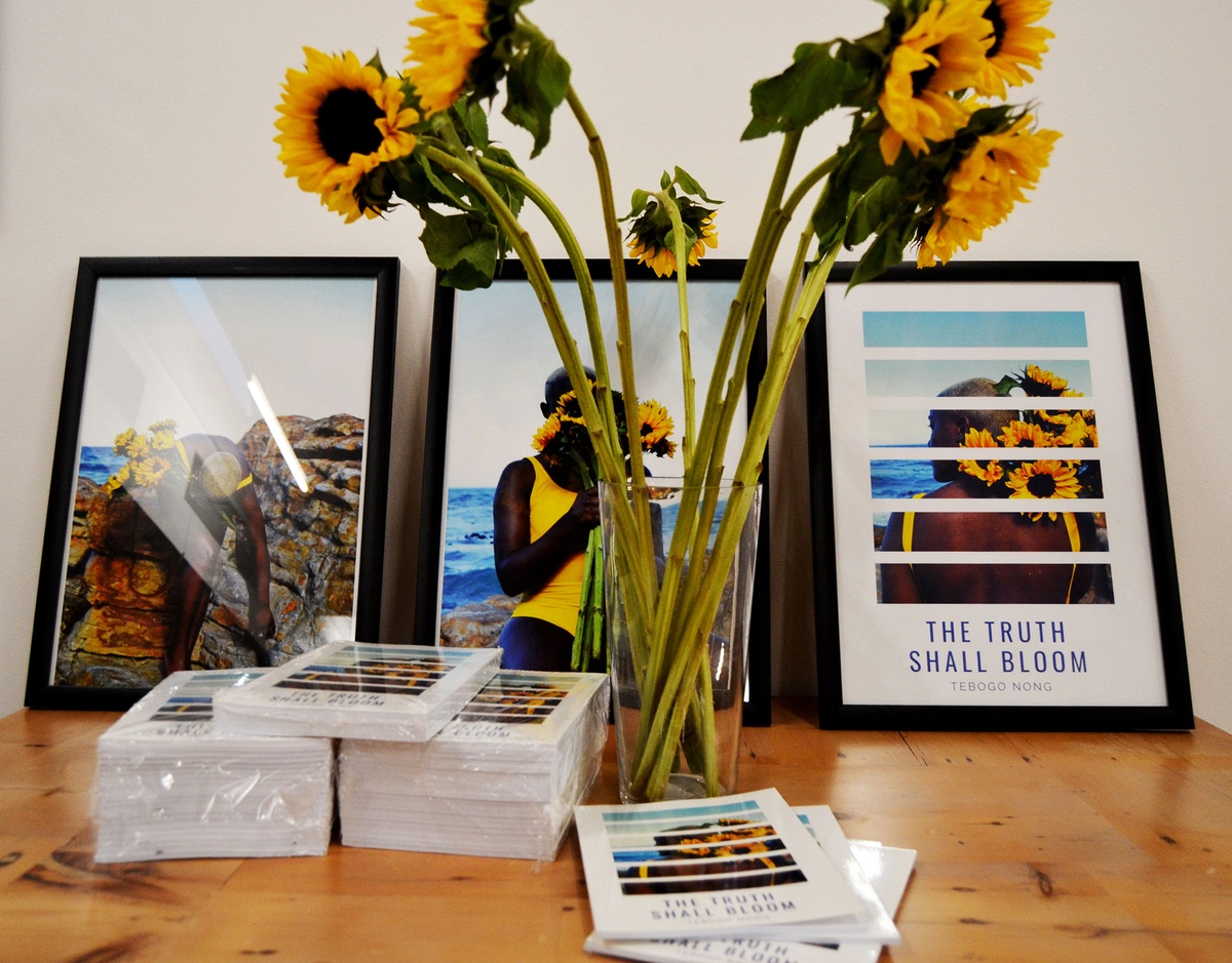 Event photograph from book launch of ‘The Truth Shall Bloom’ by Tabogo Nong on A4’s top floor that shows a closeup view of a wooden table next to a white wall. At the back, two framed photographs and a framed poster for the book lean against the wall. In the middle, a glass vase with sunflowers and a stack of flyers for the event.
