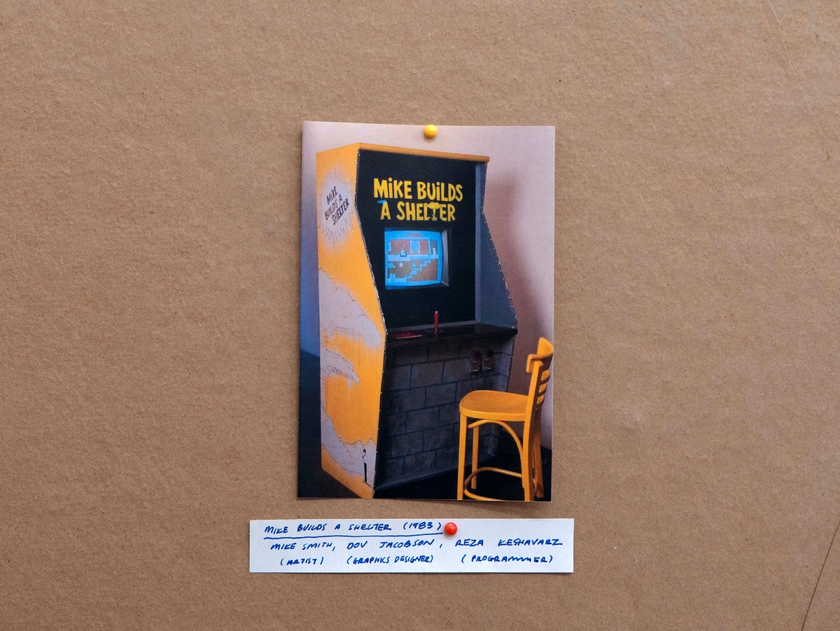 Installation photograph from Mitchell Gilbert Messina's residency in A4 Art Foundation shows a wall-mounted strip of cardboard hosting pinned research notes. The arcade cabinet as a device for interaction and animation. An image of the original _Mike Builds a Shelter_ (1983) arcade machine, considered the ‘first art videogame’. Made by artist Mike Smith, computer graphics designer Dov Jacobson, and programmer Reza Keshavarz, and developed for the exhibition _GOVERNMENT APPROVED HOME FALLOUT SHELTER AND SNACK BAR_.

