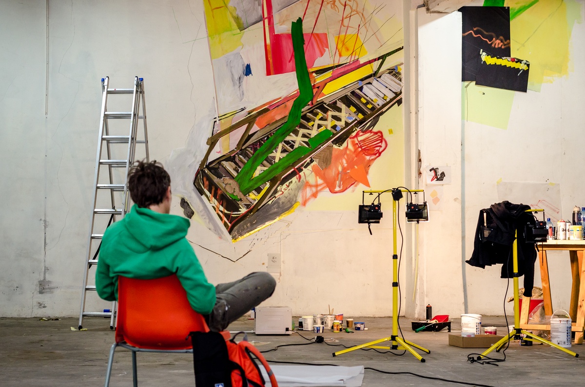 Process photograph from Dorothee Kreutzfeldt’s residency on A4’s 1st floor. At the front, Kreutzfeldt is seated on a plastic orange chair. At the back, a wall mural that features visual references to the A4’s premises on a white wall made using paper, tape, charcoal and acrylic paint.
