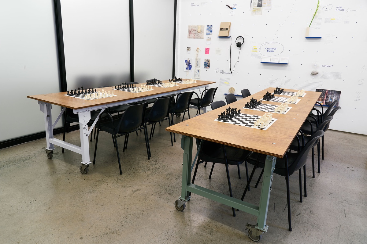 Installation photograph from the launch of Brett Seiler’s chess set in A4’s Proto museum shop that shows the chess tournament setup. Two long wooden tables with chairs host a series of travelling chess sets.
