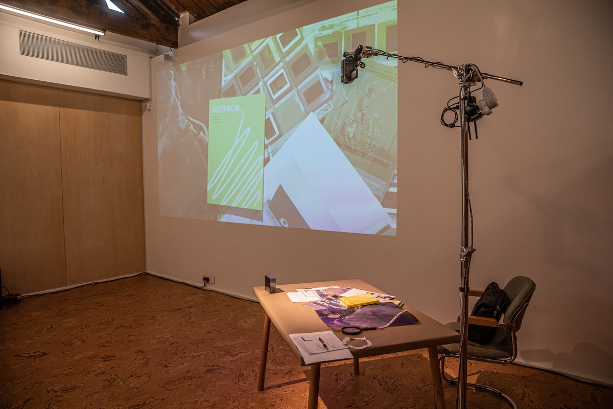 Event photograph from the book launch of 'ArtThrob: 25 Years of Art Writing in South Africa' in A4 Arts Foundation. On the right, a table for speakers. On the left, a livestream top-down view of the table is projected onto the wall.
