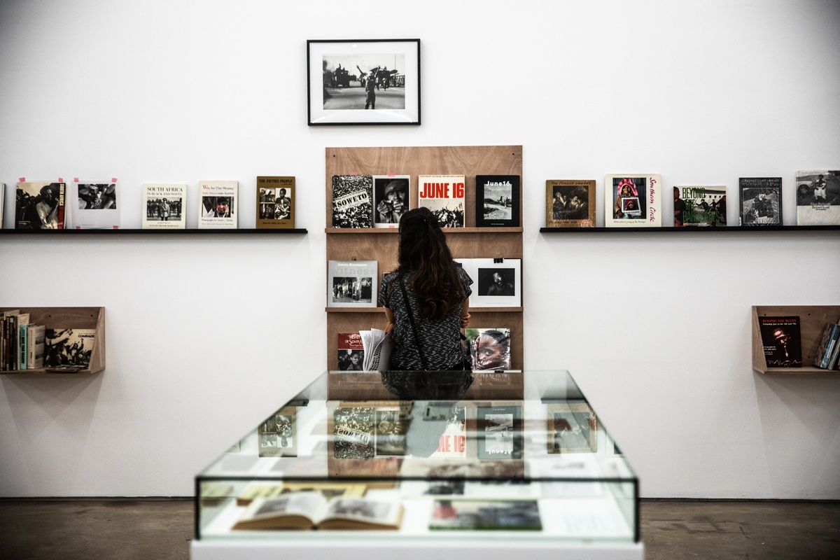 Event photograph from the opening of the Photo Book! Photo-Book! Photobook! exhibition in A4’s Gallery in an area dedicated to photobooks from the years 1967 to 1994. At the front, a glass display case. At the back, an attendee looks at a display shelf, with Peter Magubane’s photograph ‘Give peace a chance, Soweto (1976)’ mounted on the wall above it.
