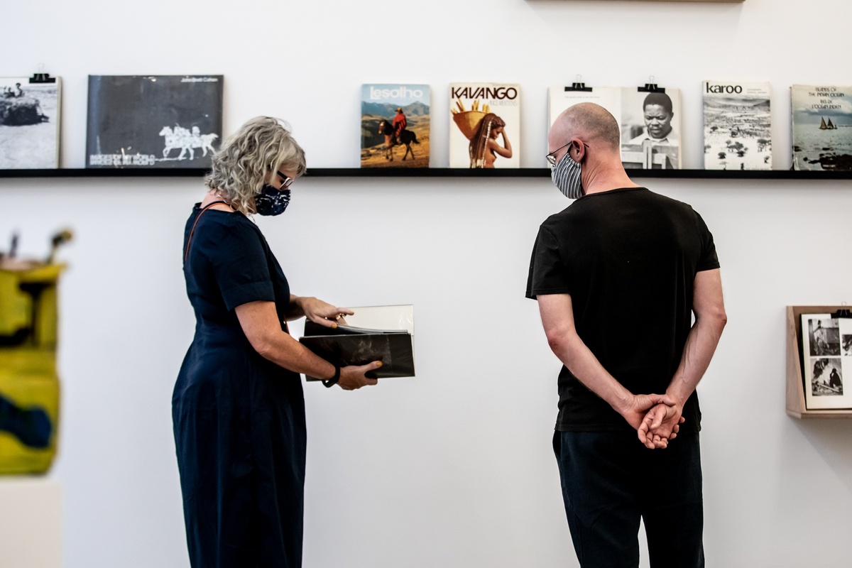 Event photograph from the opening of the Photo Book! Photo-Book! Photobook! exhibition in A4’s Gallery in an area dedicated to photobooks from the years 1967 to 1994. In the middle, two attendees stand in front of a display shelf.
