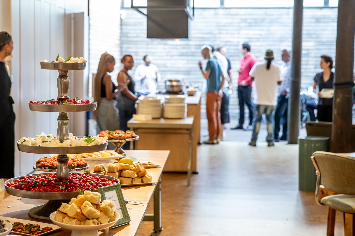 Event photograph from the preview of the 'A Little After This' exhibition in A4 Arts Foundation.   On the left, a long table and counter top are laden with food. At the back, attendees congregate on the patio.
