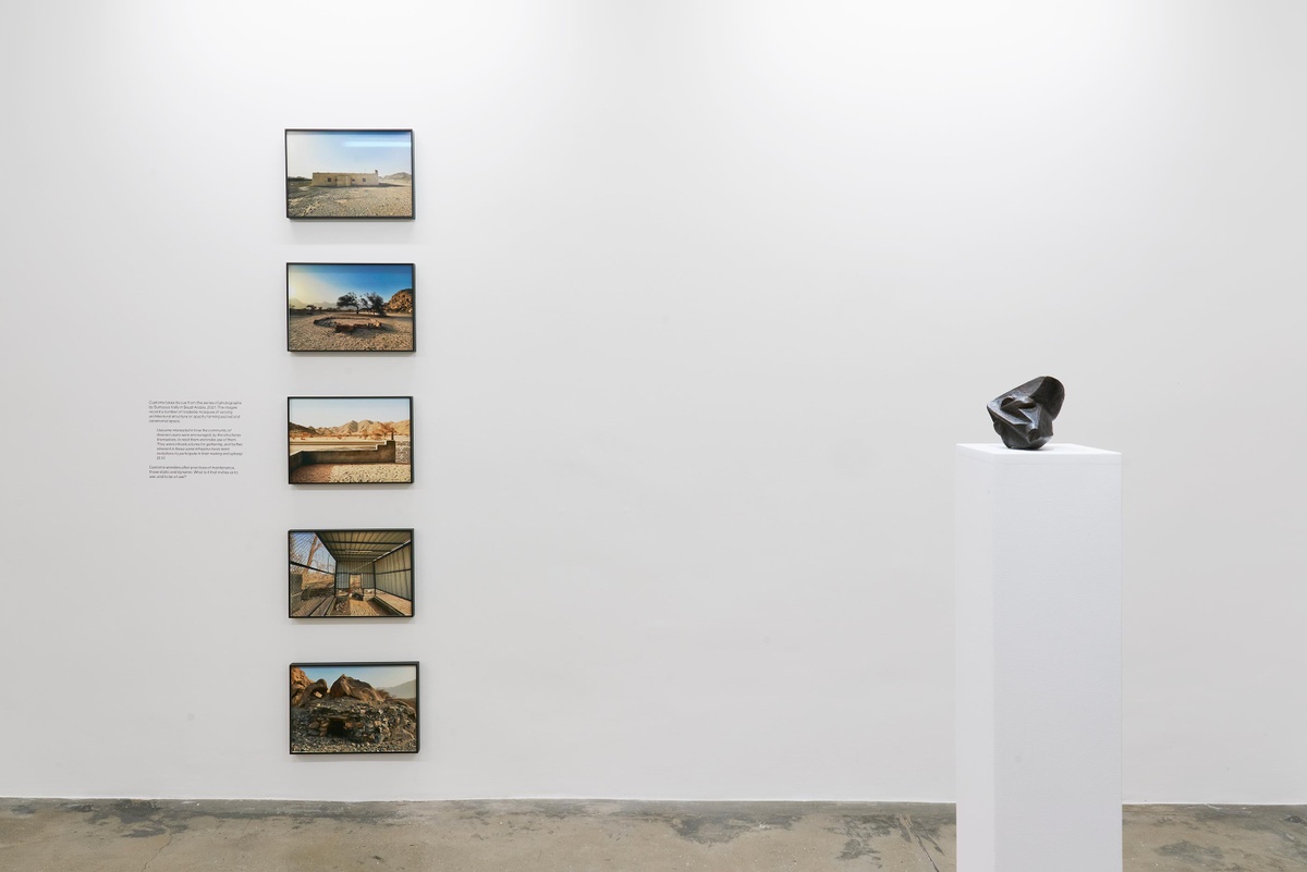 Installation photograph from the Customs exhibition in A4’s Gallery. On the left, five framed photographic prints from Sumayya Vally’s ‘Roadside Mosques’ series are mounted on a white gallery wall in a vertical column. On the right, Ezrom Legae’s bronze sculpture ‘Face’ sits on a white plinth.
