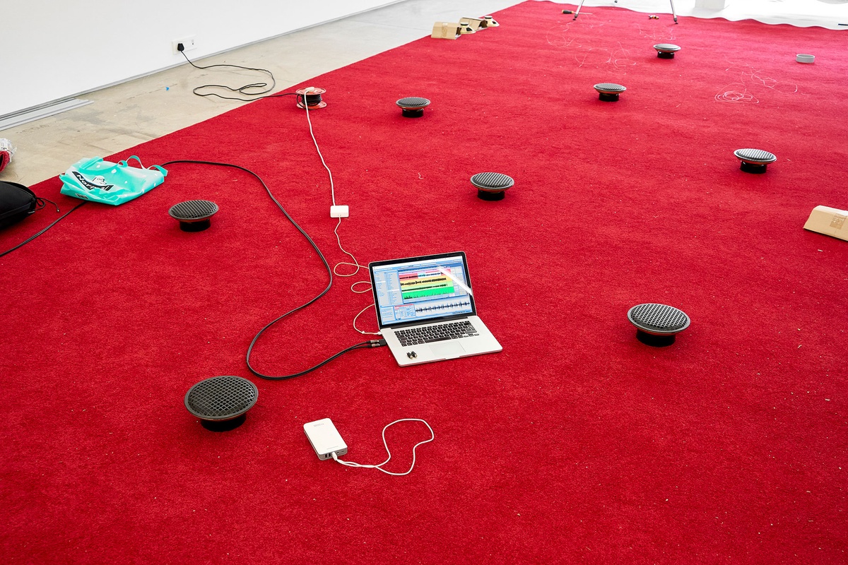 Process photograph from The Future is Behind Us exhibition in A4’s Gallery. At the front, James Webb’s ‘Prayer’ installation is being set up with a red carpet, speakers and a laptop processing audio files.
