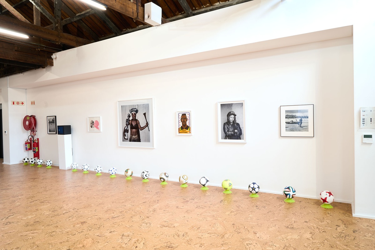 Installation photograph from the 2022 rendition of Exhibition Match on A4’s second floor. On the floor, soccer balls sit on plastic stands at the base of a white wall. On a white plinth, Robin Rhode’s animation ‘Hondtjie’ is displayed on a screen. On the wall are Andile Komanisi’s photograph ‘Soccer Indaba’, Penny Siopis’ lithograph ‘Pinky Pinky (Ronaldo)’, Pieter Hugo’s photograph ‘Good Enough Mabaso, Orlando Pirates supporter, Coca-Cola Cup semi-final, Rustenburg, 2005’, Callan Grecia’s painting ‘Lucas Radebe’ and Zanele Muholi’s photograph ‘Portia Modise, Kagiso, Krugersdorp, 2016’.
