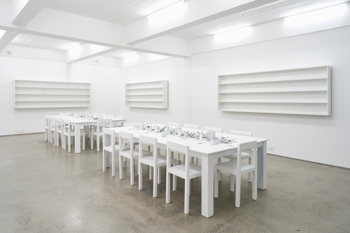 Installation photograph from the Customs exhibition in A4’s Gallery that shows Yoko Ono’s installation ‘MEND PIECE, A4 Arts Foundation, Cape Town version’ in A4’s ground-floor Reading Room. At the front, two white rectangular tables with chairs hold ceramic fragments, tape, twine and scissors. At the back, white wall-mounted shelving units line the white wall.
