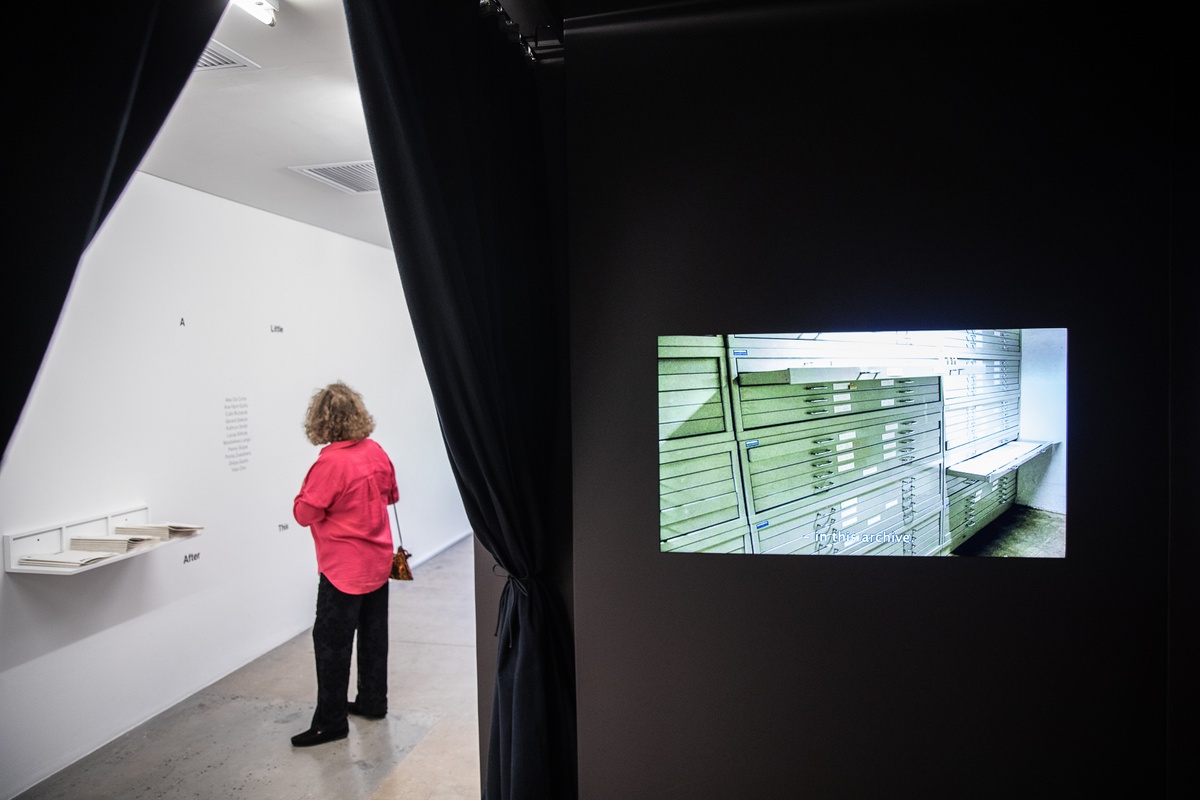 Installation photograph from the 'A Little After This' exhibition in A4 Arts Foundation's gallery. On the right, Ane Hjort Guttu's HD video 'Untitled (The City at Night)' plays on a screen in a darkened section of the gallery at the bottom of a flight of stairs. On the left, an attendee examines the title of the exhibition described on the wall in vinyl.
