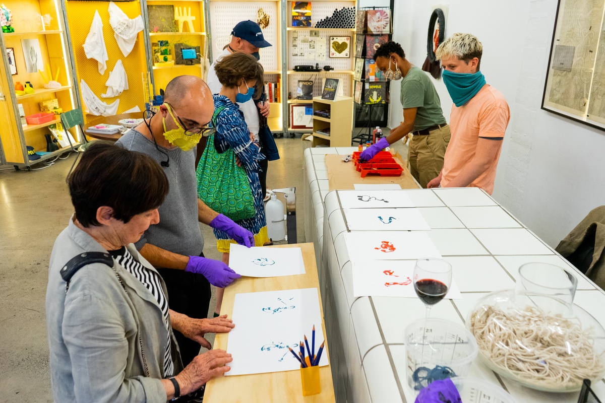 Event photograph from ‘Knots’, a conversation between Igshaan Adams and Josh Ginsburg, marking the close of ‘Open Production’, Adams’ hybrid studio/exhibition in A4’s Gallery. At the reception desk, A4 staff members facilitate an interactive exchange where attendees can create artworks by using threads dipped in dye as a drawing medium.
