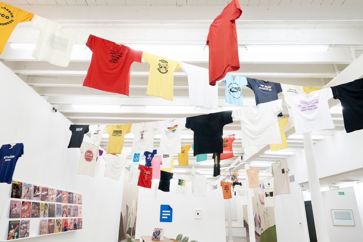 Installation photograph of the Common exhibition. Above, T-shirts with slogans from the GALA Queer Archive and SAHA are suspended on criss-crossing lines. On the left, vinyl record covers from ‘The Library of Things We Forgot to Remember’ by Kudzanai Chiurai are arranged on a slanted white shelf. At the back is ‘Google Docs’ by Hanna Noor Mahomed, a painting of the Google Docs logo.
