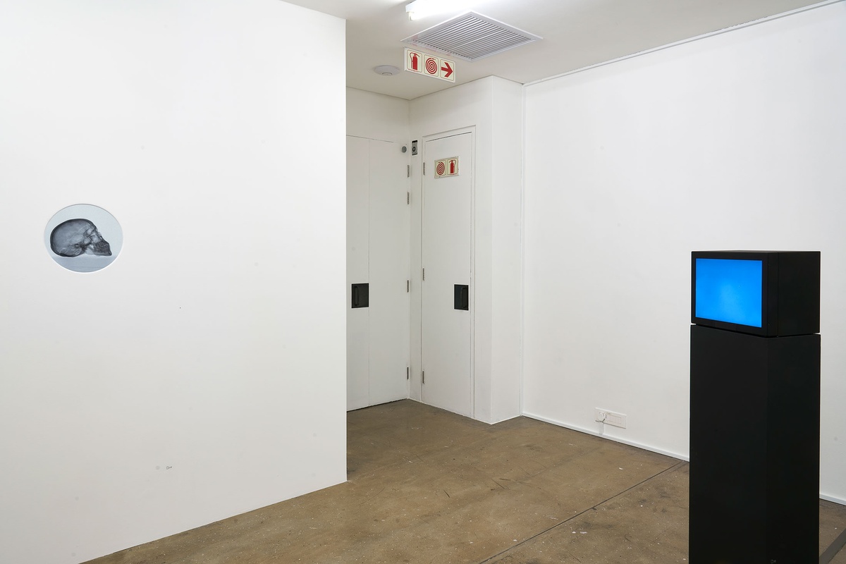 Installation photograph from the 'A Little After This' exhibition in A4 Arts Foundation's gallery. On the left, Kathryn Smith's digital animation 'The Phantom Interlocutor' playing on a screen embedded in a white wall, visible through a circular porthole. On the right, Yoko Ono's closed-circuit 'Sky T.V.' live streaming on a screen mounted on a black plinth.
