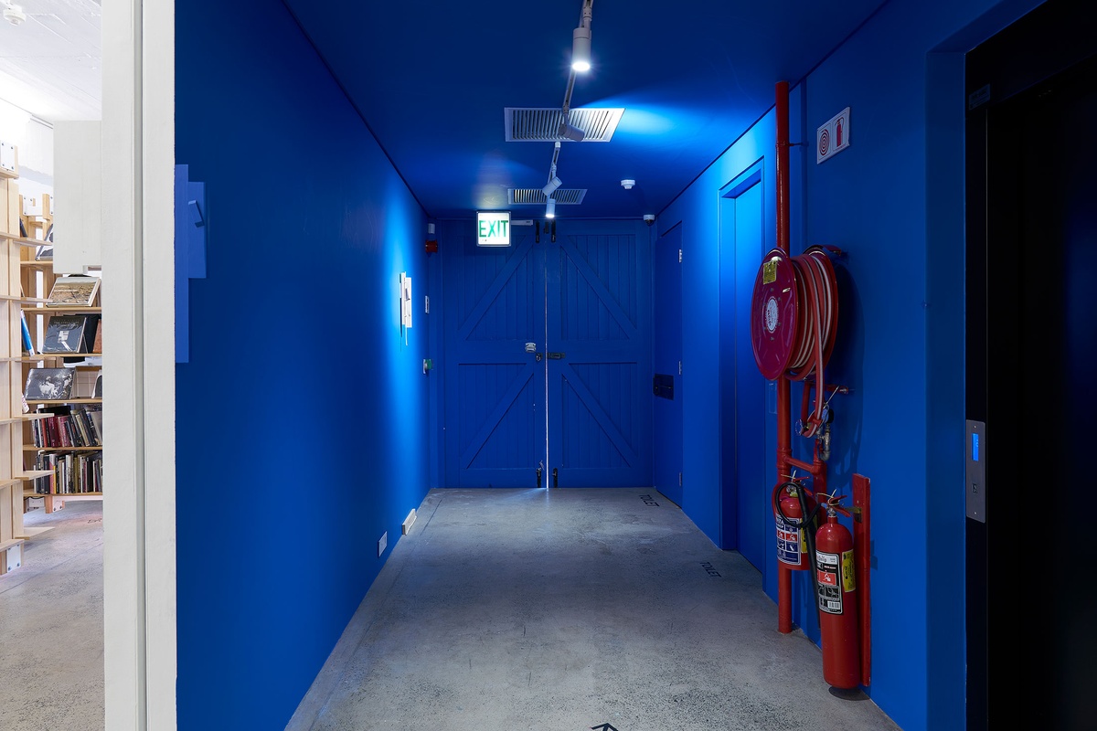 Installation photograph from Guy Simpson's Passage exhibition in A4 Arts Foundation's Goods space shows two sculptures that recall the mezuzah attached to the wall. At the front, one sculpture is painted blue to match the wall. At the back, another sculpture is unpainted, with an unpainted wooden detail at the base of wall.
