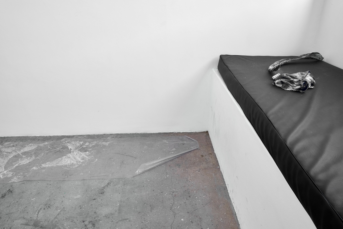 Installation photograph from the ‘without a clear discernible image’ exhibition in A4’s Gallery that depicts Kevin Beasley’s untitled installation. On the left, a sheet of heat-forming plastic sits on the gallery floor. On the right, resin hardened seaweed and fabric lie on a gallery bench.
