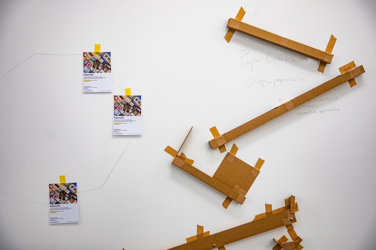 Event photograph from the opening of the Papertrails exhibition in A4’s Reading Room. On the left, flyers for the exhibition are pasted to the wall. On the right, a cardboard Rube Goldberg machine is mounted on the wall.
