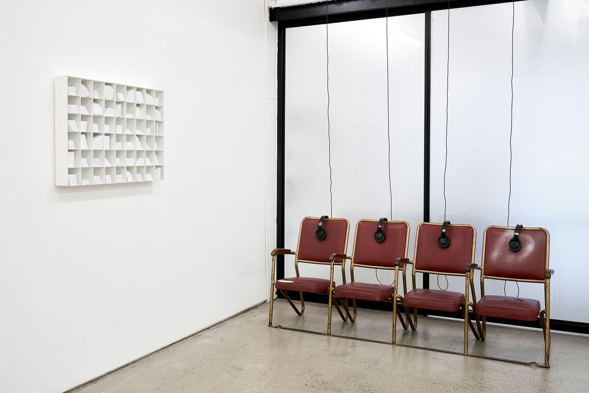 Installation photograph from the ‘Model’ exhibition in A4’s Reading Room. On the left, Kyle Morland’s paper sculpture ‘Library of Forms’ is mounted on the wall. On the right, Bhavisha Panchia’s sound work ‘Imagine you’re in a museum: What do you hear?’ features four chairs with headphones.
