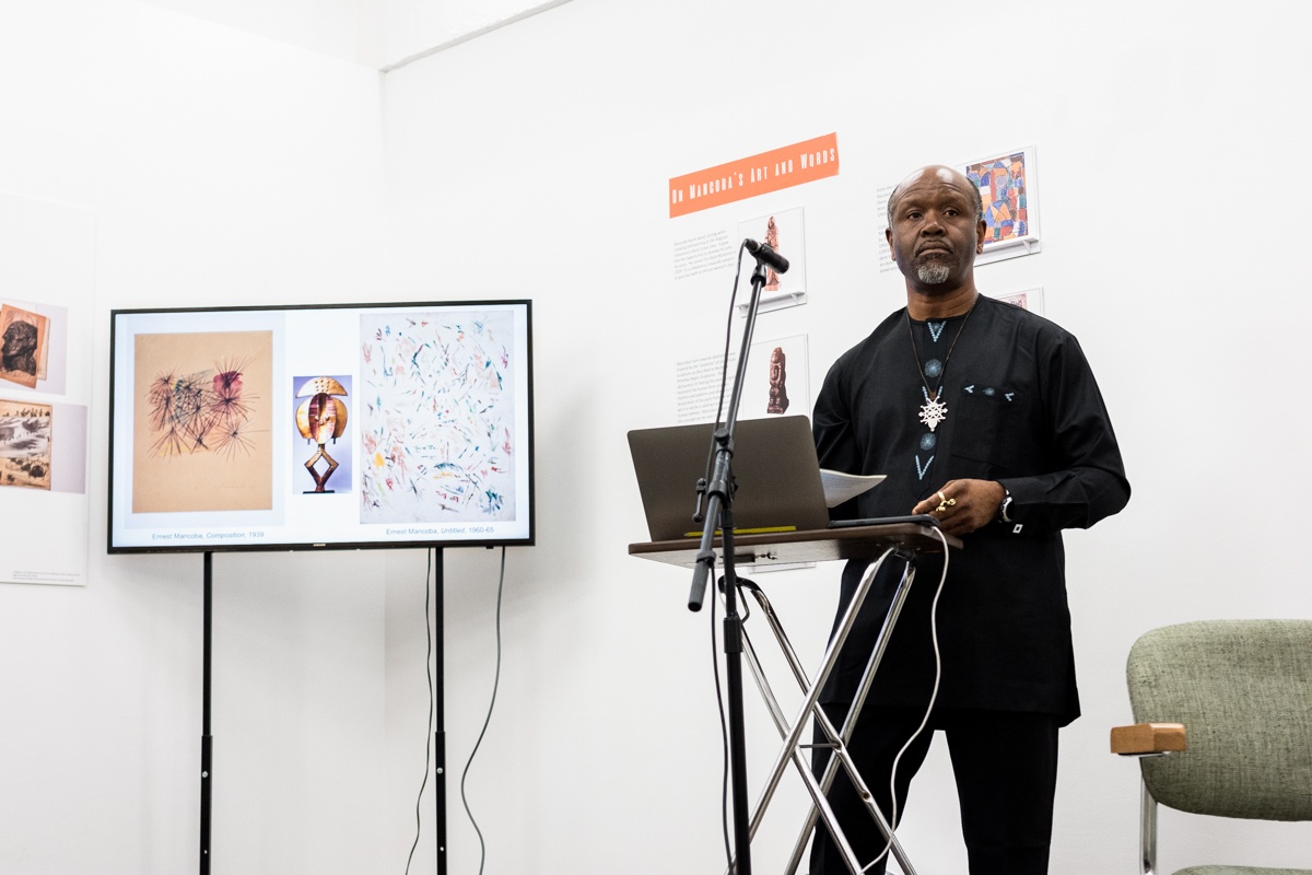 Event photograph from the Ernest Mancoba Symposium at A4 Arts Foundation. On the right, Prof Chika Okeke-Agulu stands at a podium with a laptop and microphone. On the left, a television screen displays images of artworks by Mancoba.
