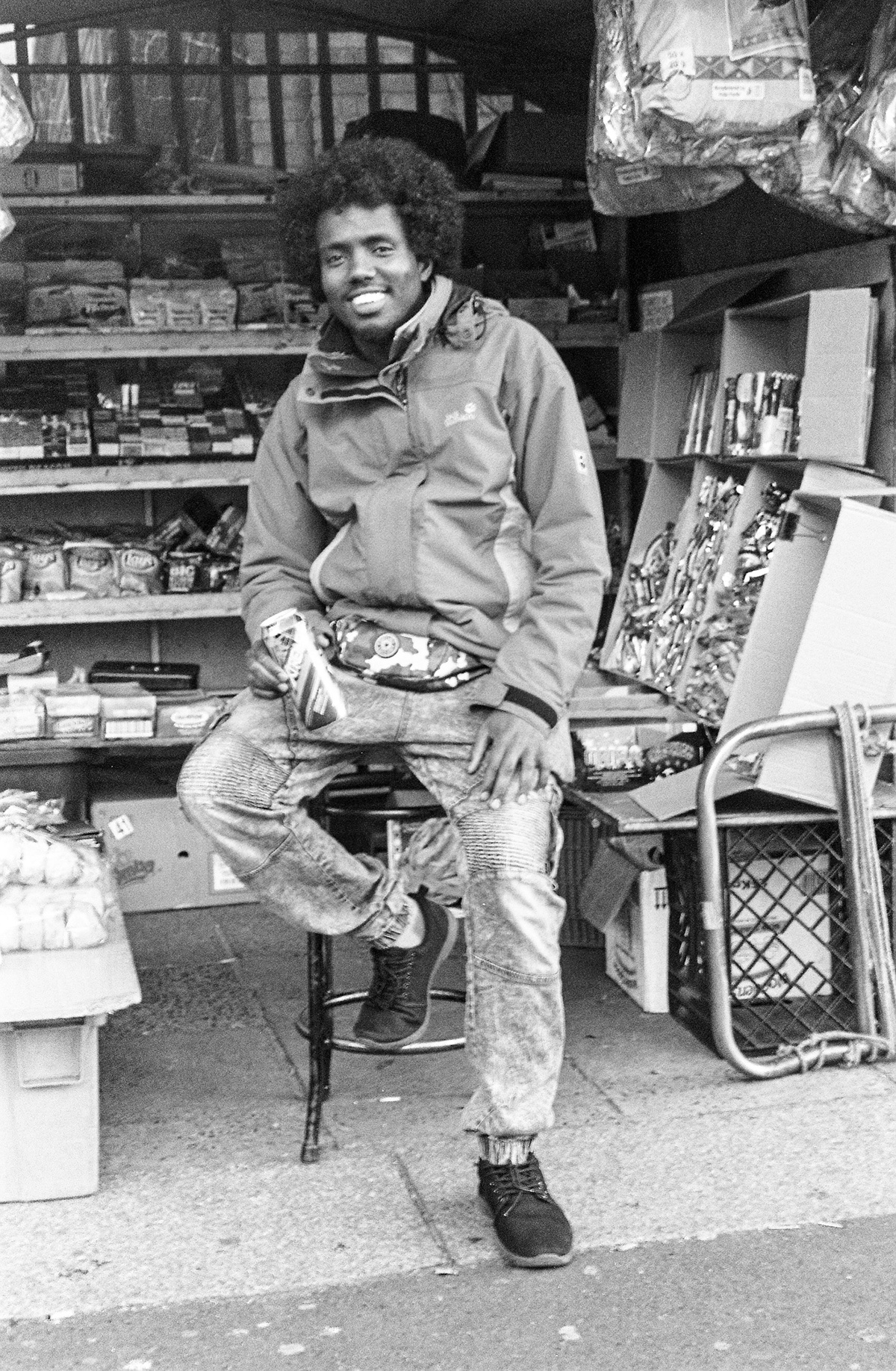 Monochrome event photograph from ‘Contact’, a collaboration between A4’s proto~ museum shop and Oath Magazine, taken by a participant. A smiling shopkeeper sits on a chair in front of makeshift shelves.
