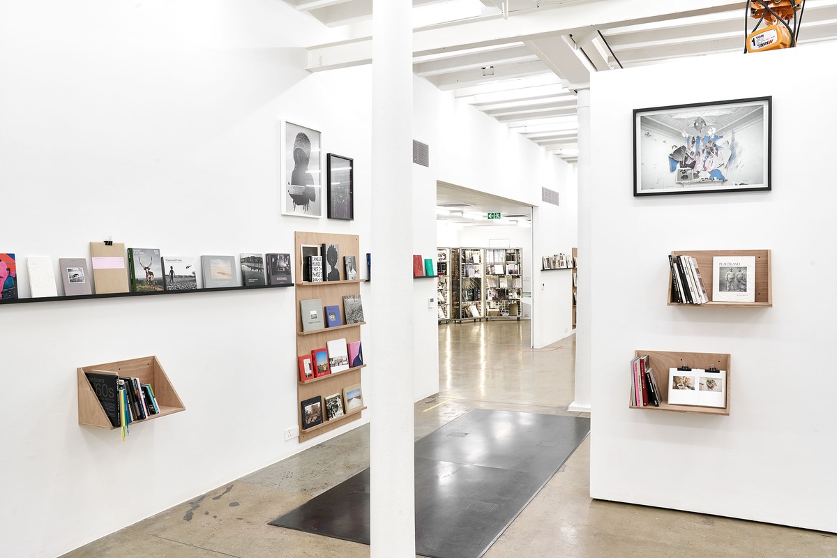 Installation photograph from the Photo Book! Photo-Book! Photobook! exhibition in A4’s Gallery in an area dedicated to photobooks from the years 1994 to 2022. On the left, Zanele Muholi’s photographs ‘Hlanzeka III, Vineyard Hotel, Room 153, Cape Town, 2017’ and ‘Bangizwenkosi, The Sails, Durban’ are mounted above a wooden display shelf. On the right, Zander Blom’s photograph ‘Les Demoiselles d'Avignon. Bedroom 1, Corner 3, 7.00 pm., Tuesday, 7 August 2007’ is mounted above a wooden display shelf.
