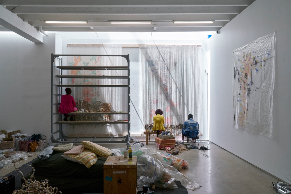 Process photograph from ‘Open Production’, Igshaan Adams’ hybrid studio/exhibition in A4’s Gallery. At the back, three of Adam’s studio assistants are weaving with threads suspended on looms attached to the top of the gallery walls. At the back of the threads tracings made onto tracing paper are used as guides. On the lefthand side, one of the assistants is seated on metal scaffolding.
