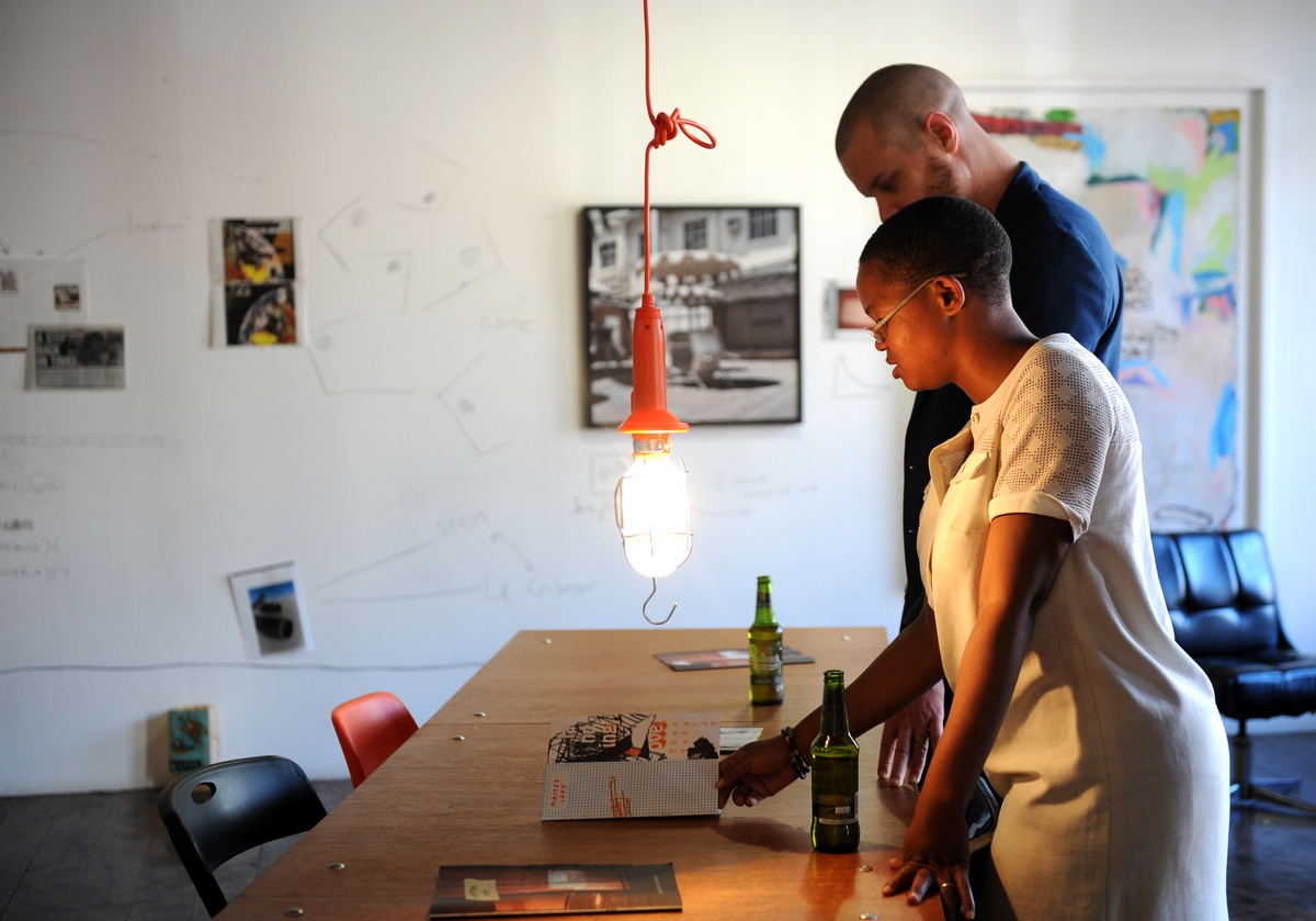 Event photograph from the offsite ‘Office Politics’ exchange. At the front, two attendees interact with copies of Tabogo George Mahashe and francis burger’s ‘Camera Obscura #3’ publication and Andrew Juries and Lauren Theunissen’s ‘Have a nice Day’ publication a wooden table.
