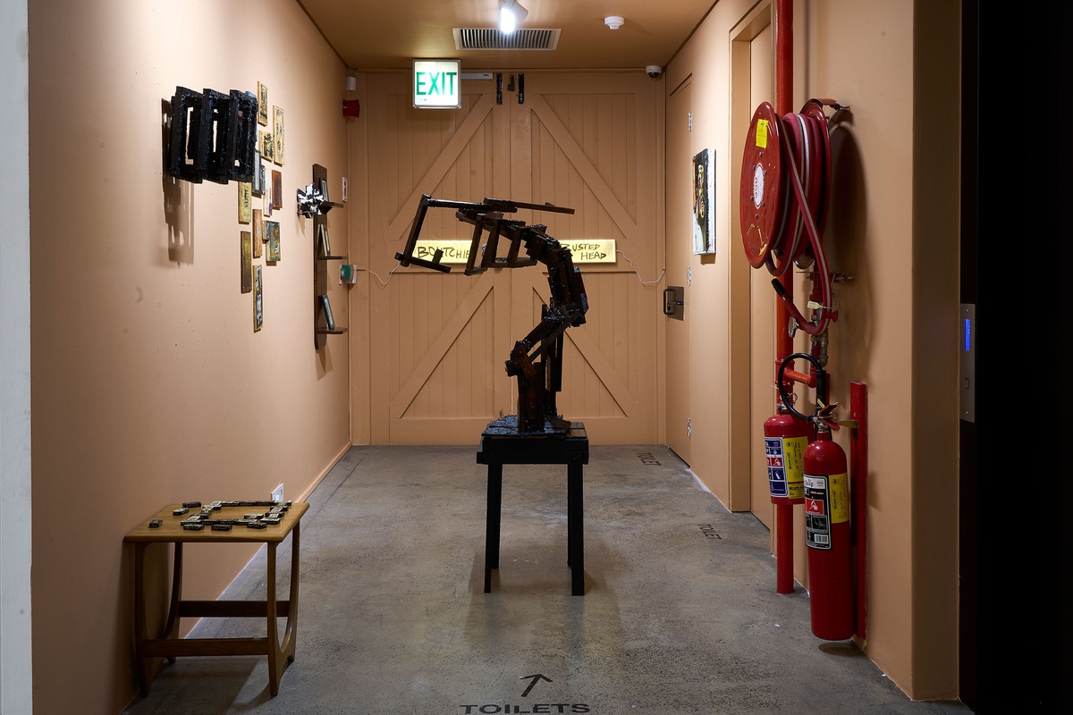 Installation photograph from Phillip Newman's exhibition in A4 Arts Foundation's Goods space. In the middle, a wooden sculpture stands on the concrete floor. On the left, drawings on small wooden panels are arranged on the wall. At the back, a wooden shelf holds framed drawings.
