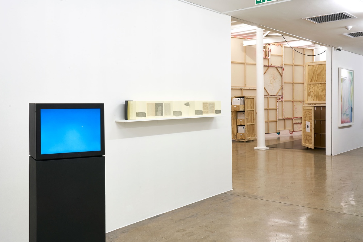 Installation photograph from the 'A Little After This' exhibition in A4 Arts Foundation's gallery. On the left, Yoko Ono's closed-circuit 'Sky T.V.' live streaming on a screen mounted on a black plinth. On the right, Colin Richards' Moleskin, collage and ink work 'A Little After This' is displayed on a wall-mounted shelf.
