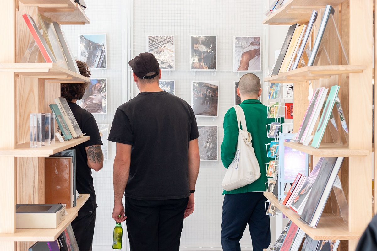 Event photograph from the book launch of CJ Chandler's 'twist of a knee' in A4 Arts Foundation's Proto museum store. At the back, photographic prints from the book are mounted on a metal wall-mounted grille. In the middle, attendees are looking at the display. At the front, two of Proto's wooden bookshelves frame the image on the left and right.
