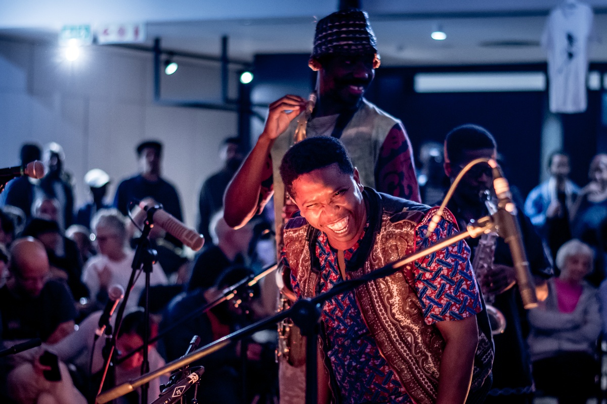 Event photograph from a performance by Shabaka and the Ancestors, with special guest Bra Louis Moholo-Moholo, on A4’s top floor. At the front, three musicians performing with microphone stands and wind instruments. At the back, attendees are seated and standing.

