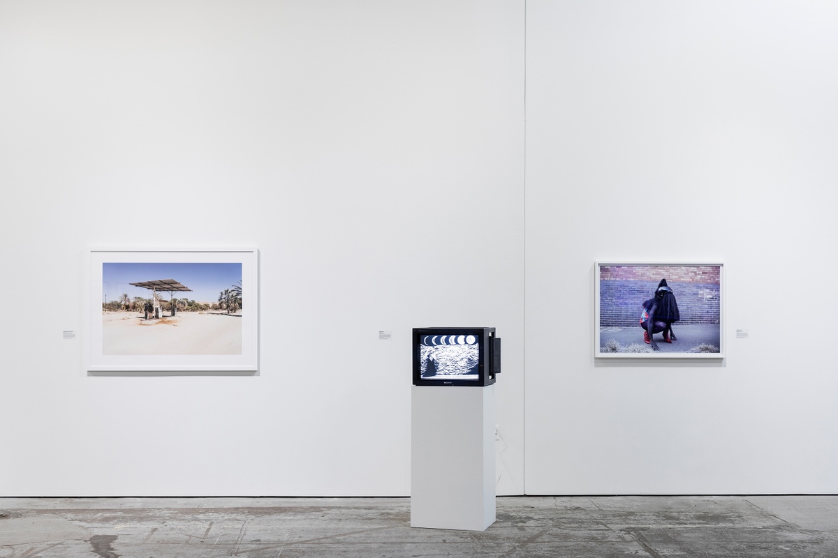 Installation photograph from ‘Crossing Night: Regional Identities x Global Context’ exhibition at the Museum of Contemporary Art Detroit. On the left, Margaret Courtney Clarke’s photograph ‘Petrol Pump, Sesfontein, Namibia’ is mounted on the wall. In the middle, Robin Rhode’s video ‘The Moon is Asleep’ is displayed on a screen sitting on a plinth. On the right, Athi-Patra Riga’s photograph ‘The Naivety of Beiruth’ is mounted on the gallery wall.
