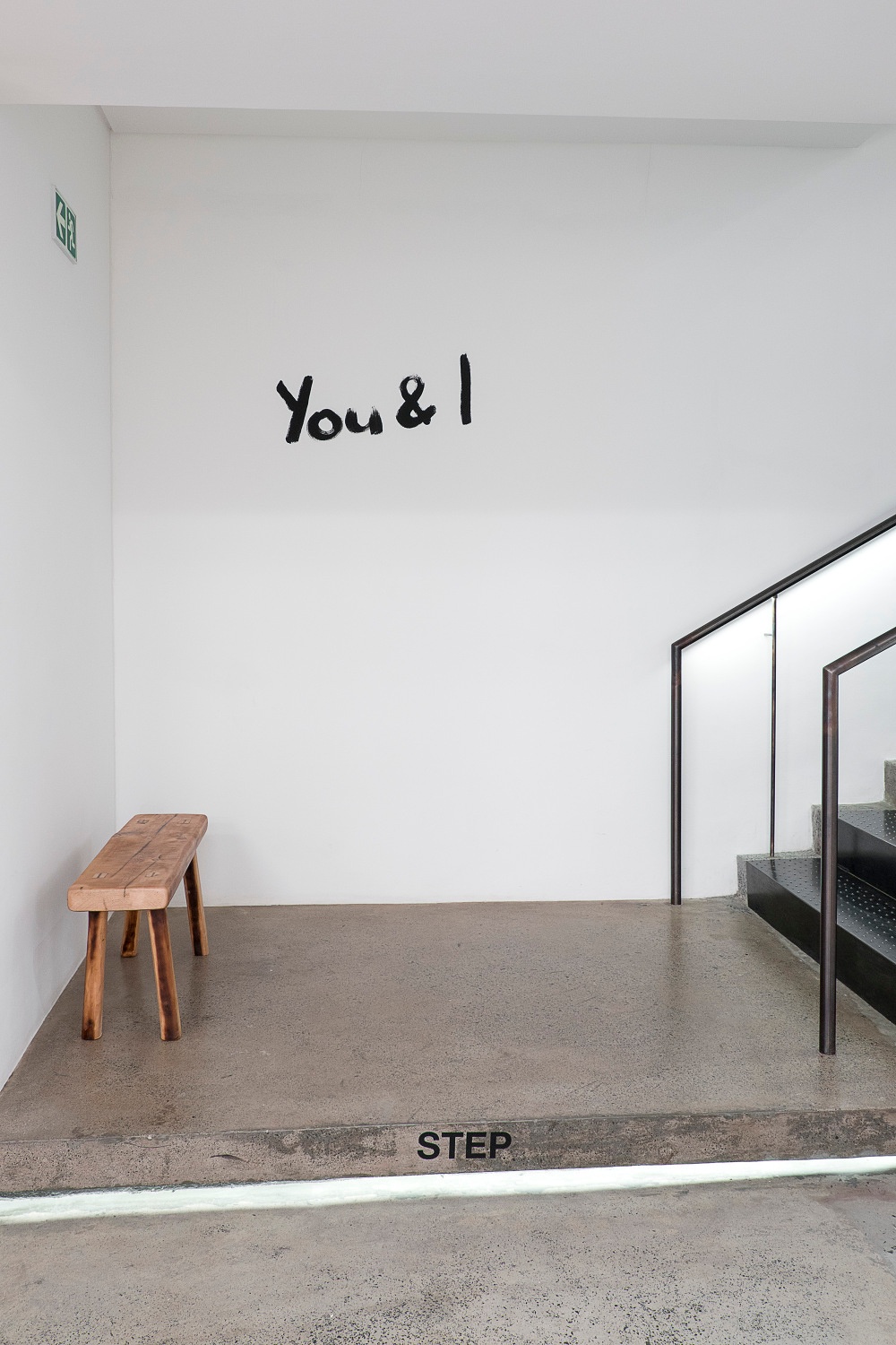 Installation photograph from the You & I exhibition in A4’s Gallery. On the ground floor, the exhibition title ‘You & I’ is painted on a wall at the base of the stairway in black paint.
