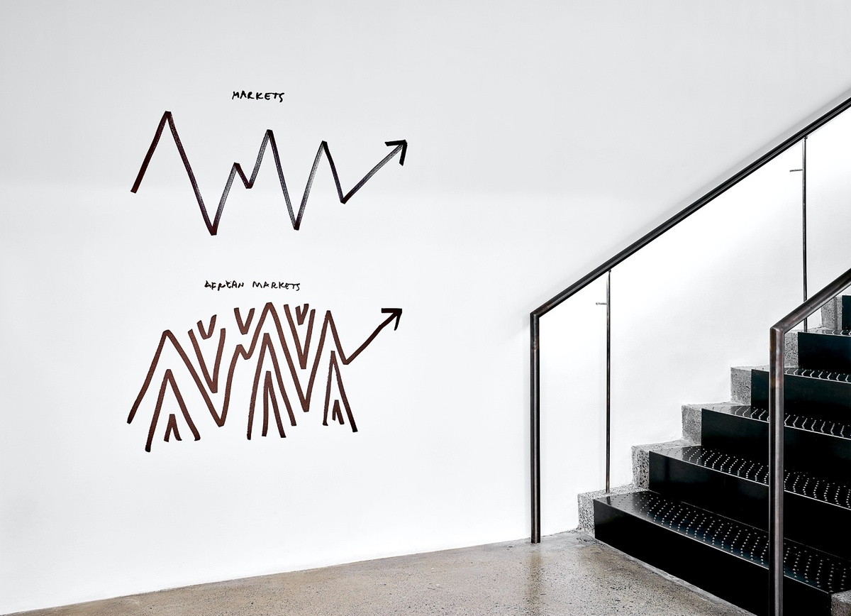 Installation photograph from Dan Perjovschi’s ‘The Black and White Cape Town Report’ exhibition in A4’s Gallery that shows black felt pen marker drawings on white walls at the base of A4’s staircase. At the top, a zig zagging line that ends in an arrow pointing up the stairs with the phrase ‘Markets’. At the bottom, the figure is repeated with meany additional line sections above and below it and the phrase ‘African Markets’.
