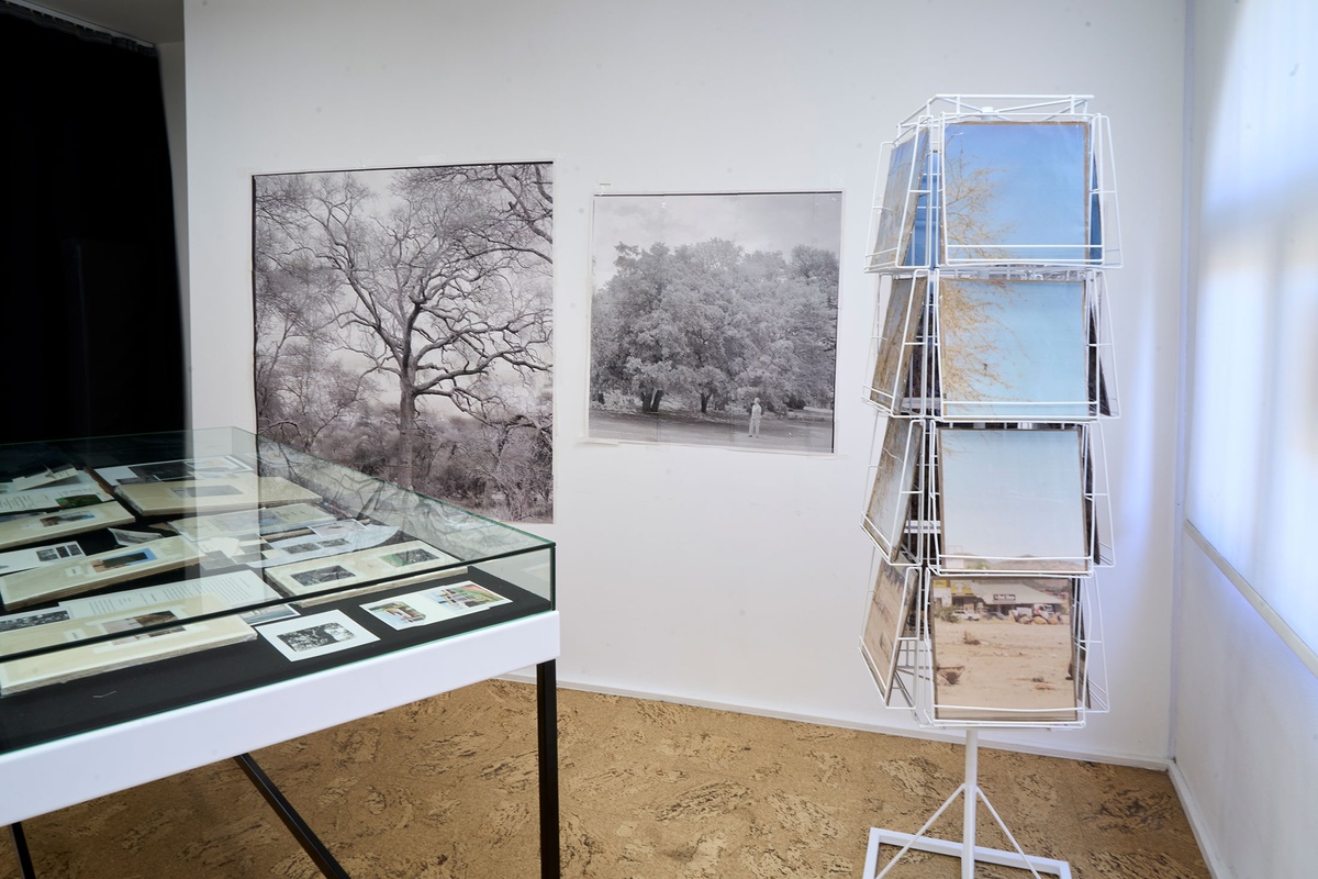 Installation photograph of Michael Tymbios’ studio during his residency in A4 Arts Foundation. On the left, a freestanding display case holds photographs and ephemera. On the right, a freestanding rotating record display stand holds photographic prints. At the back, photographic prints of various sizes are mounted on a white wall.
