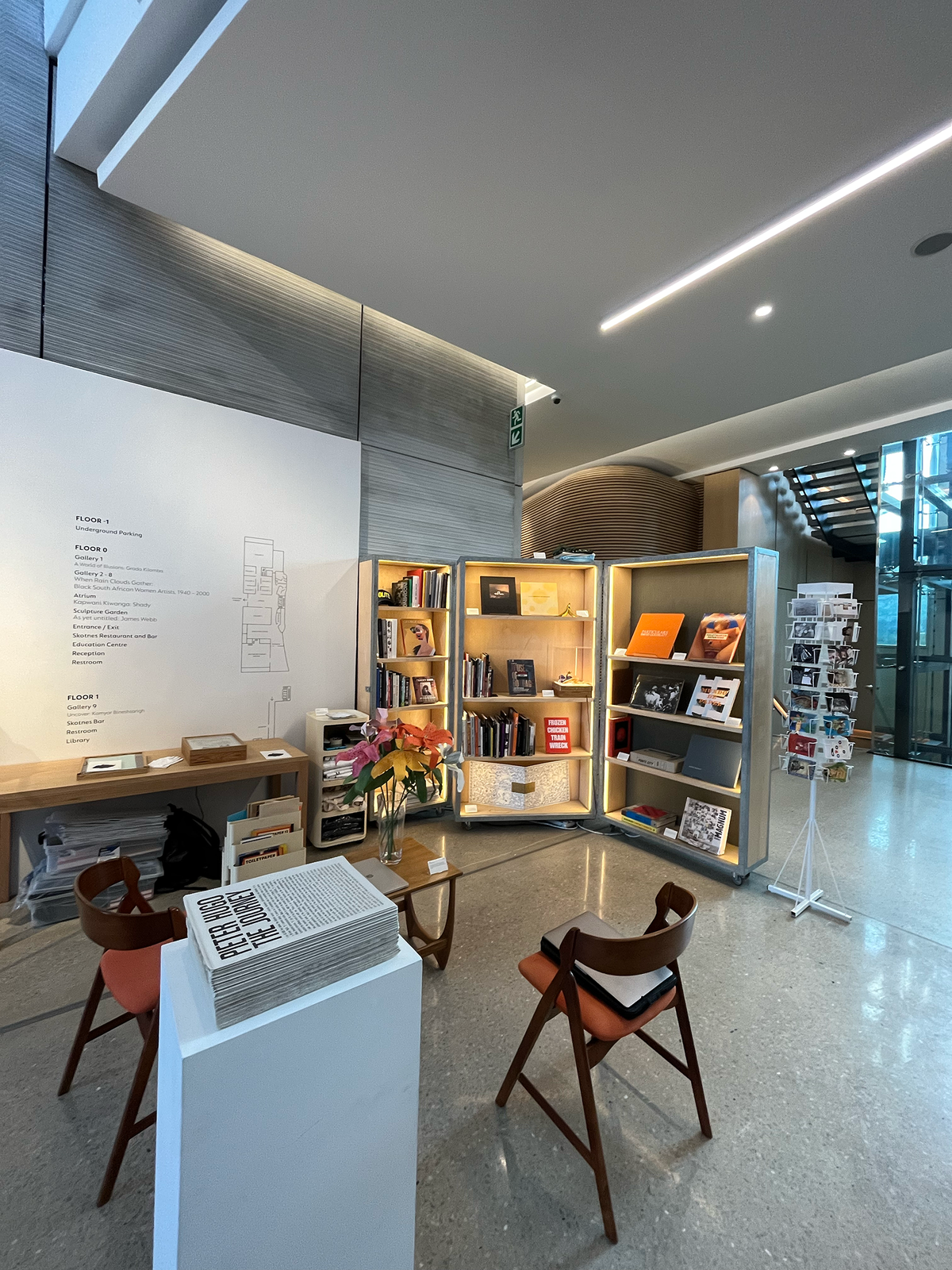 Installation photograph of Proto's booth at the 2022 Cape Town Art Book Fair. At the back, a wood and metal display cabinet holds a selection of books. At the front, a white plinth features a stack of Pieter Hugo's newsprint publication 'The Journey'.
