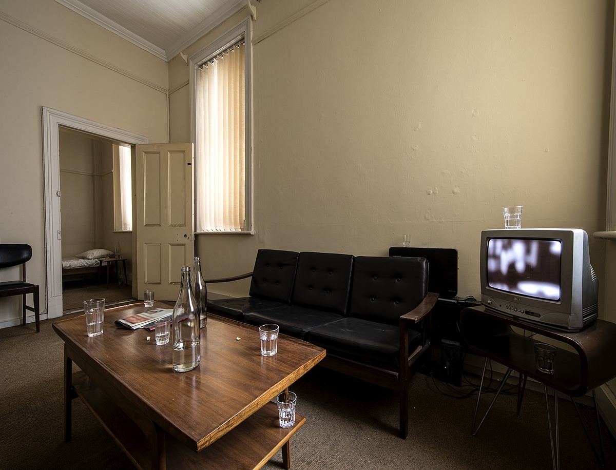 Installation photograph from Gain Maria Tosatti’s offsite exhibition ‘My Hart is so Leeg soon ’n Spieël (My Heart is a Void, the Void is a Mirror)’. On the right, a television set on a table. In the middle, a black leather couch. On the left, a two-tiered wooden table with printed matter, glass bottles, drinking glasses and teeth.
