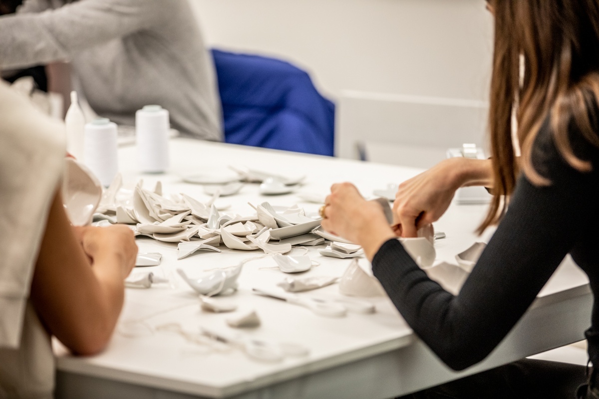 Event photograph from the opening of the Customs exhibition in A4’s Gallery that shows Yoko Ono’s installation ‘MEND PIECE, A4 Arts Foundation, Cape Town version’ in A4’s ground-floor Reading Room. A close-up view shows participants constructing sculptural forms using ceramic fragments, twine, tape and scissors on a white table.
