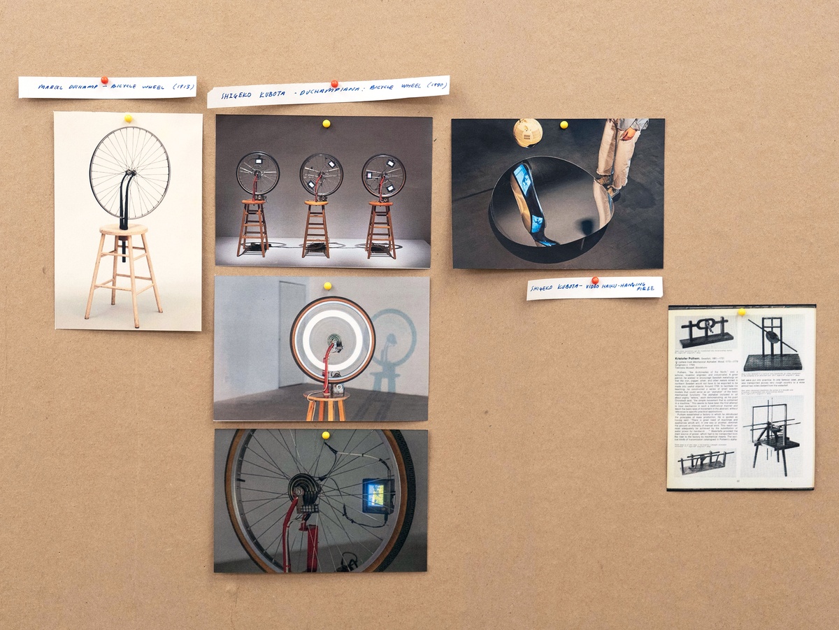 Installation photograph from Mitchell Gilbert Messina's residency in A4 Art Foundation shows a wall-mounted strip of cardboard hosting pinned research notes. Images of (1) Marcel Duchamp’s _Bicycle Wheel_, 1913 alongside (2) Shigeko Kubota’s _Duchampiana: Bicycle Wheel One_, 1990 a single-channel video, (3) Shigeko Kubota’s _Video Haiku–Hanging Piece_, 1981 and (4) Kristofer Polhem’s _Letters from Mechanical Alphabet_, 1772-1779.
