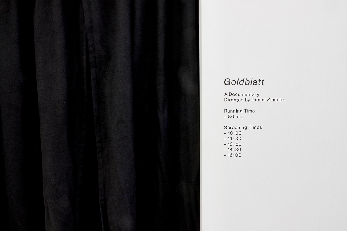 Installation photograph from the ‘Picture Theory’ exhibition in A4’s Gallery. On the left, a black curtain covers the video room entrance. On the right, a wall vinyl reads ‘Goldblatt: A Documentary Directed by Daniel Zimbler’.
