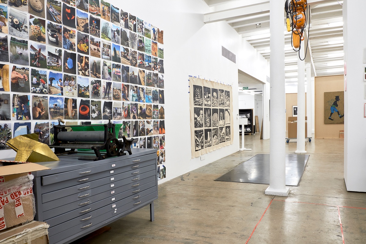 Installation photograph from the 2018 rendition of ‘Parallel Play’ in A4’s Gallery. On the left, a set of metal drawers with a manual metal printing roller on top of it, with rows of photographic prints from Jo Ratcliffe’s archive on the wall behind it. In the middle, a large sheet of paper with drawings by Jonah Sack.
