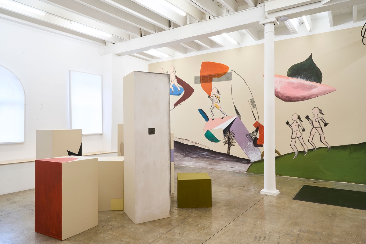 Installation photograph of Common exhibition, featuring an installation by Hanna Noor Mahomed called ‘The Prodigal Daughter.’ On the left, a group of plinths painted with geometrical shapes and objects to resemble a city. On the right at the back, a wall mural with five femme figures running among various objects and geometrical shapes.
