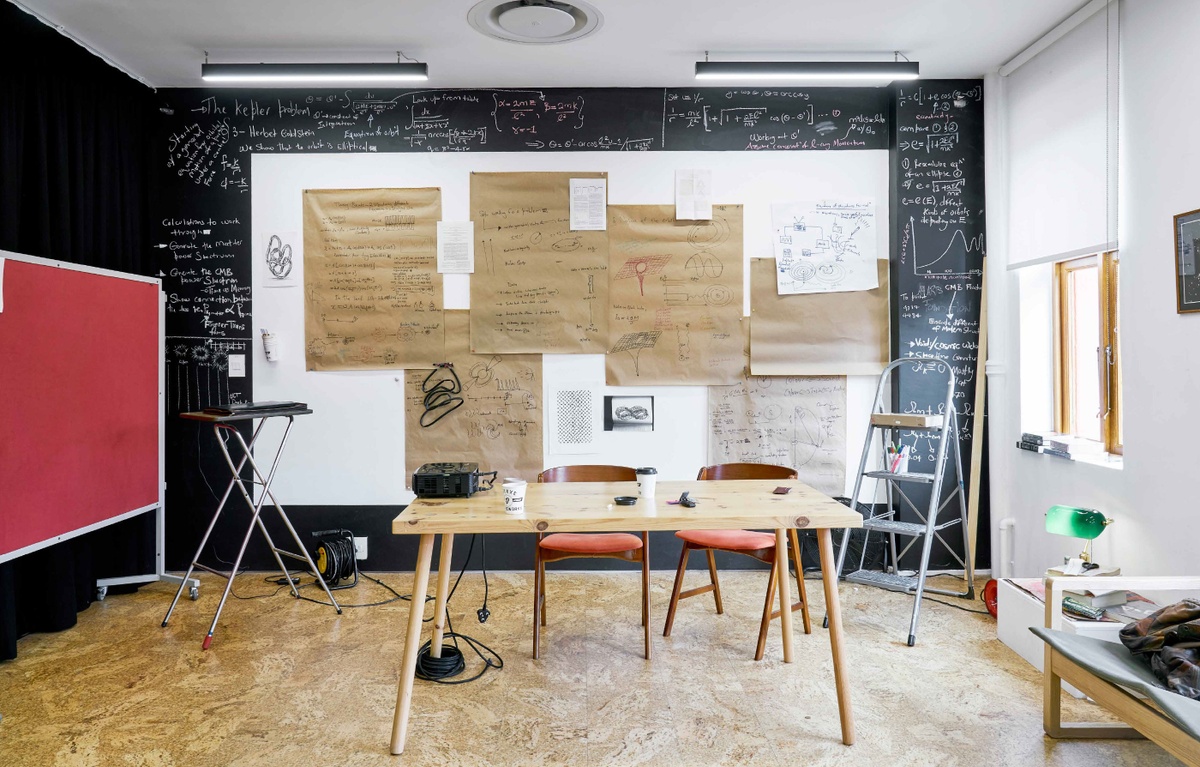 Process photograph from Sukuma Mkhize’s residency on A4’s top floor. At the back, the wall of A4’s studio is covered in sheets of annotated brown paper and diagrams and notes in chalk. At the front, a wooden table hosts a projector and paper coffee cups.
