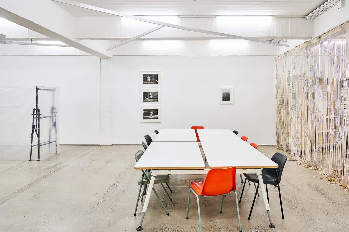 Installation photograph from the ‘Nowhere - No we're - Now here’ exhibition on A4’s ground floor. On the left, Gian Maria Tosatti’s mixed media installation ‘Void #2’ sits on the floor. In the middle, Sabelo Mlangeni’s photographic series ‘Invisible Woman’ and Santu Mofokeng’s photograph ‘End of the Line, KZ2-Auschwitz (Landscapes of Trauma)’ are mounted on the gallery wall. On the right, Igshaan Adams’ mixed media installation ‘Al asma ul Husna’ is mounted to the ceiling. At the front, laminate tables with seating.
