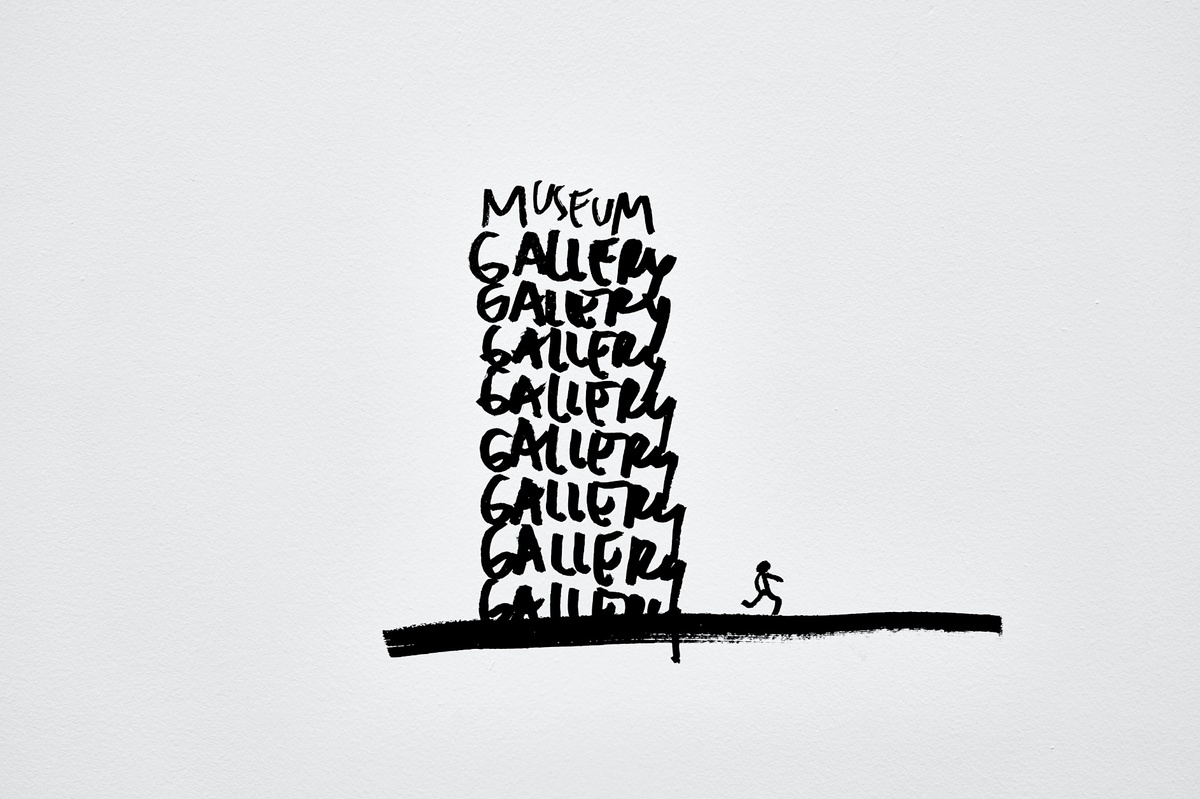 Installation photograph from Dan Perjovschi’s ‘The Black and White Cape Town Report’ exhibition in A4’s Gallery that shows black felt pen marker drawings on white walls. A large thick line is topped with eight repetitions of the phrase ‘gallery’ and one of ‘museum’ at the top, with a small stick figure running away from the edifice of repetition.
