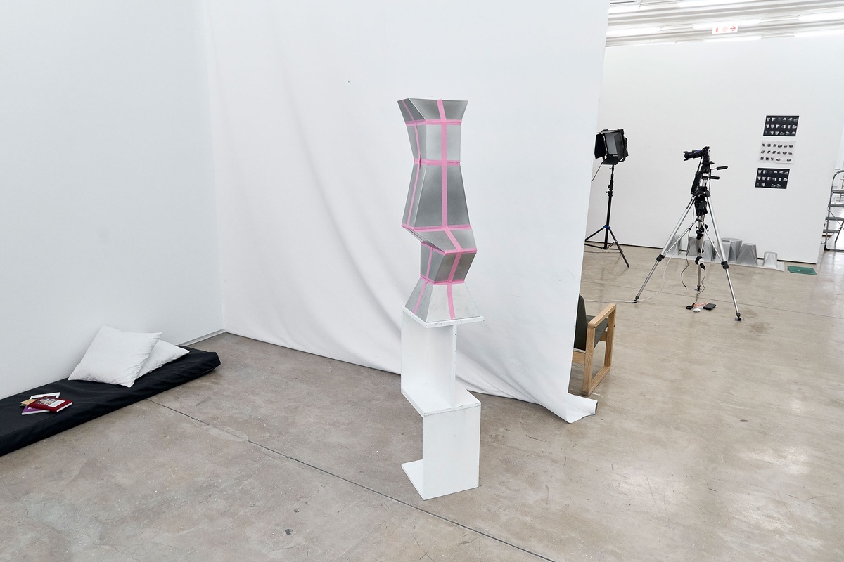 Process photograph of Kyle Morland’s working space during the 2nd rendition of ‘Parallel Play’ in A4’s Gallery. In the middle, Morland’s aluminium sculpture ‘Stacked Facets’ is overlaid with pink tape and sits on a custom shaped plinth. On the left, a mattress with pillows lays on the floor.
